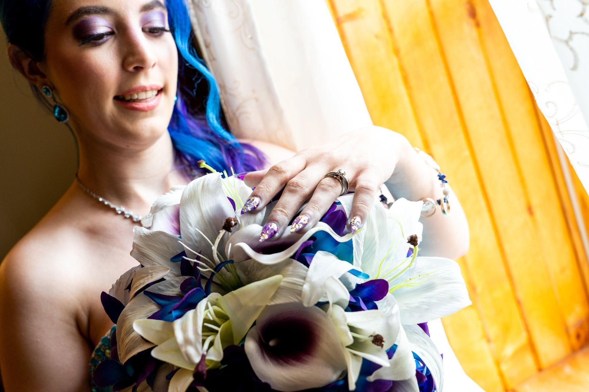 Bride with blue-streaked hair and floral bouquet, wearing jewelry, smiling near a window with warm light.