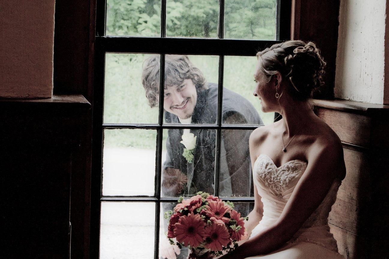 Groom peeks through window at bride in wedding dress, holding flowers. Smiling, intimate moment.
