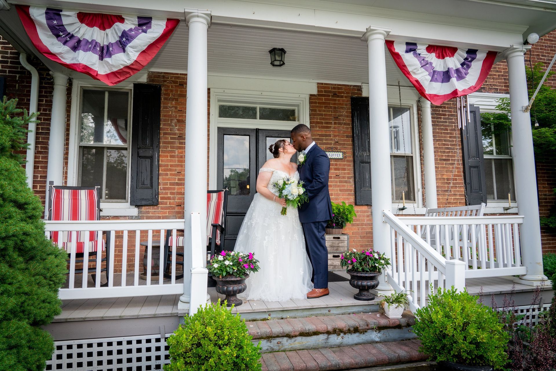Couple kissing on porch of brick house, decorated with flags and flowers.