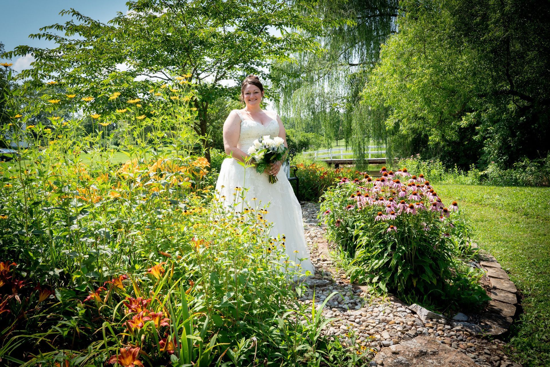 Bride in white dress holding bouquet, standing in a sunny flower garden.