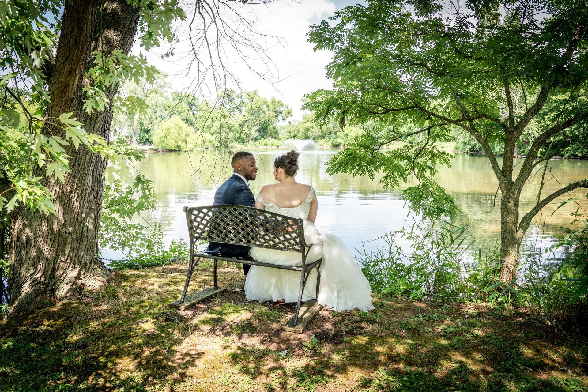 Newlyweds sit on a bench by a lake, surrounded by trees, sunny day.
