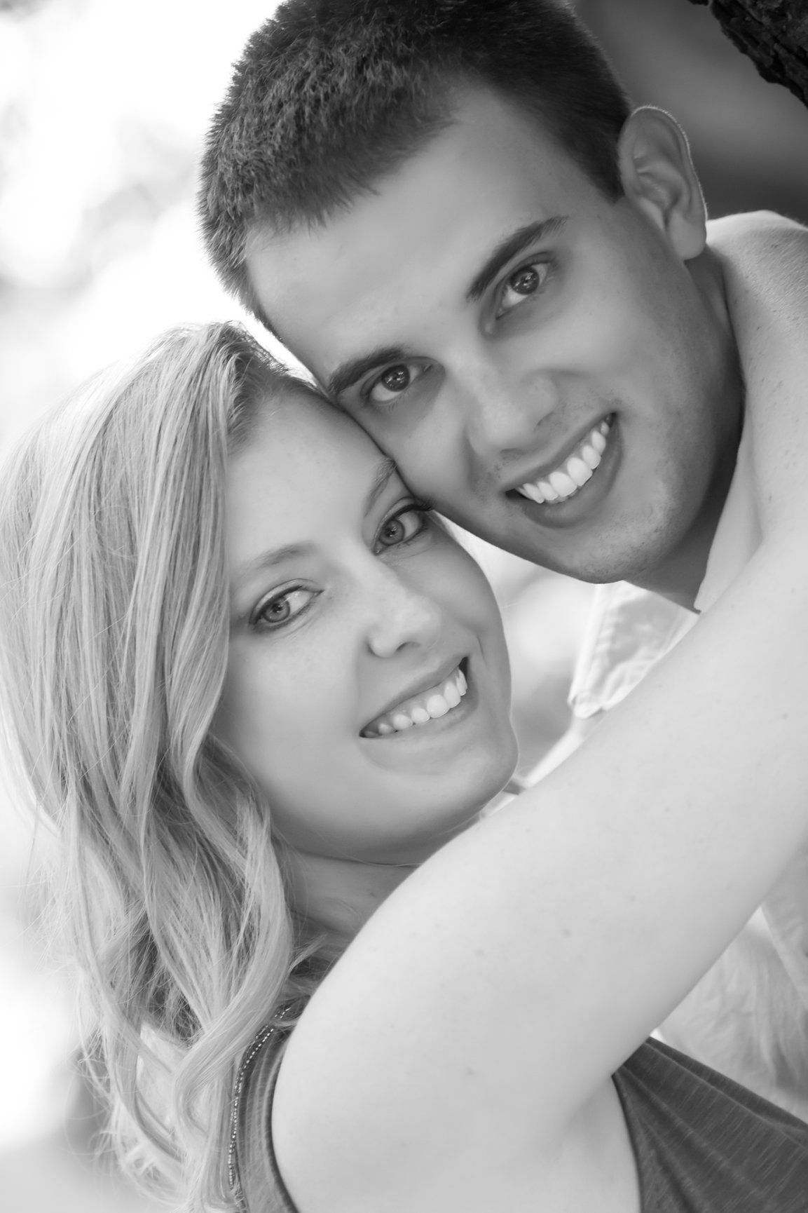 Smiling couple embracing, woman in foreground, man behind her, both looking at camera.
