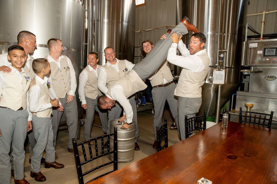 Groomsmen pose in brewery; one doing handstand on keg, others laughing. Neutral colors, indoor setting.