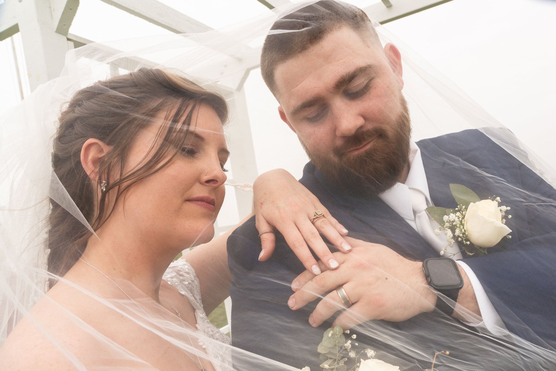 Bride and groom embracing, under a veil. The bride's hand is on the groom's. They are looking down.