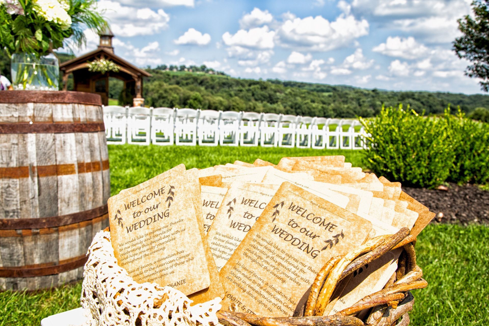 Wedding programs in a basket, outdoor ceremony setting with white chairs, barrel, and scenic view.