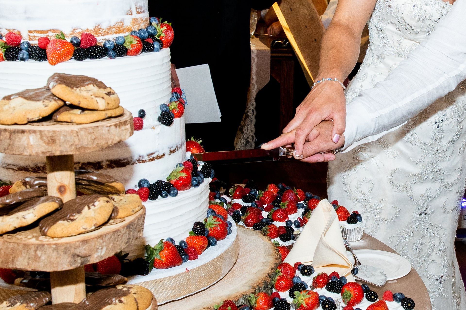 Couple cutting a tiered wedding cake decorated with berries and cookies.