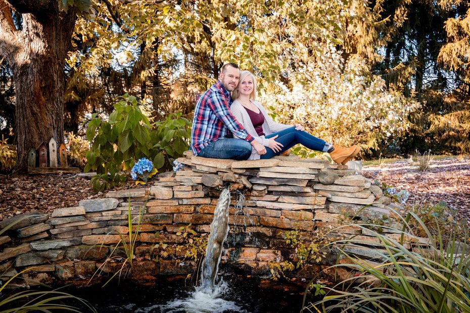 Couple sits on stone ledge near waterfall, fall foliage in background. Man in plaid, woman in jeans smiles.