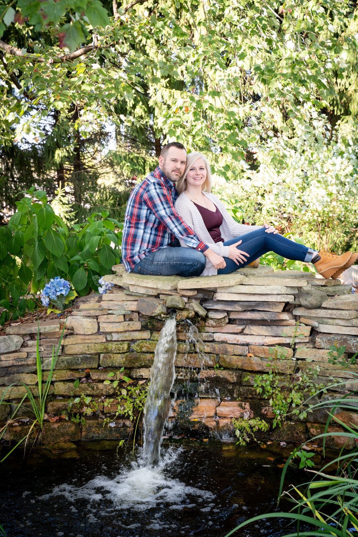 Couple seated on a stone wall bridge by a small waterfall, smiling.