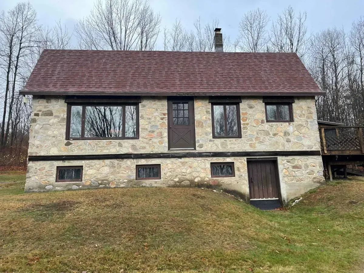Small stone cottage with a red roof on a grassy slope, set against leafless trees.