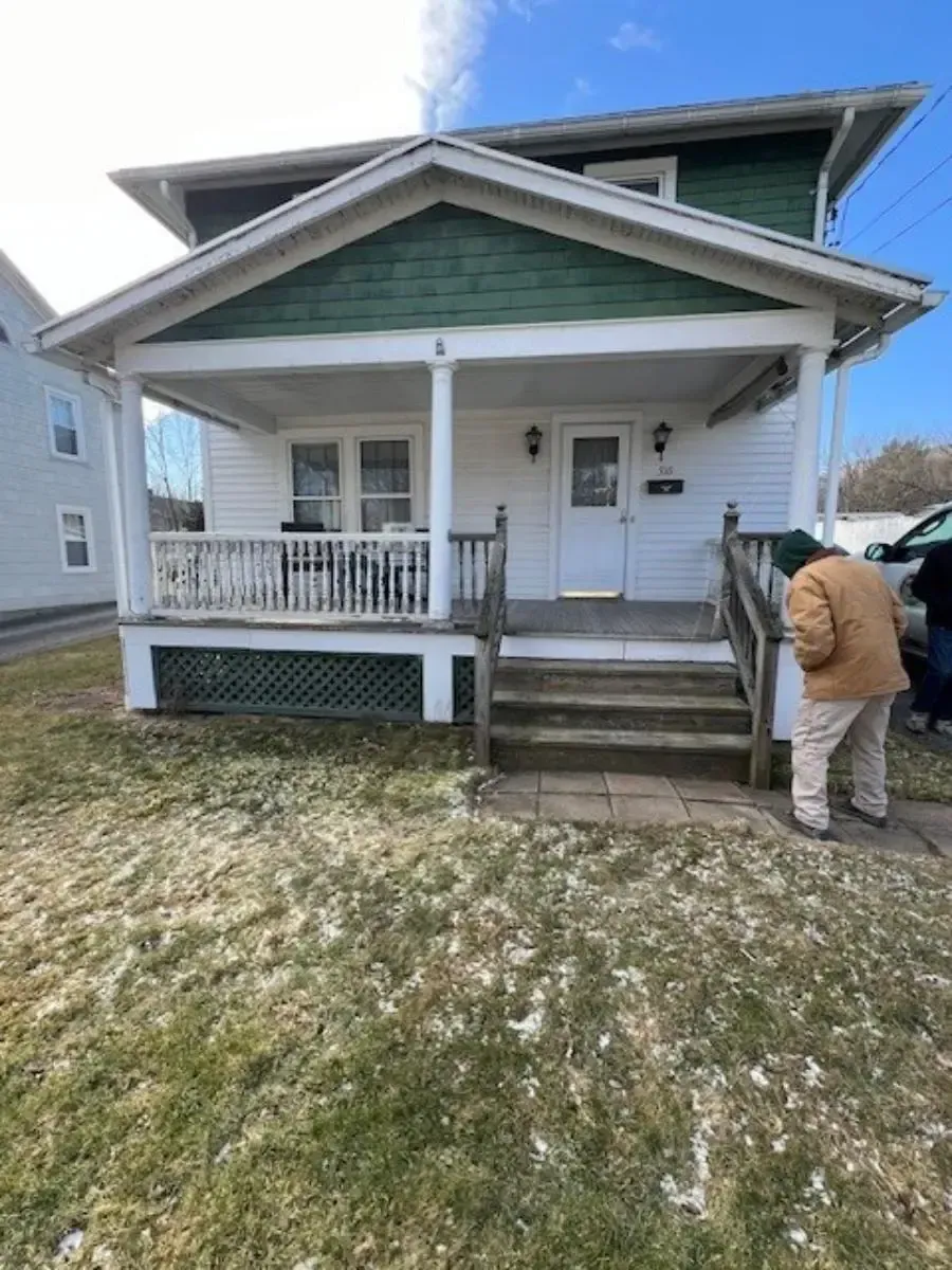 A man is standing in front of a white house with a green roof.
