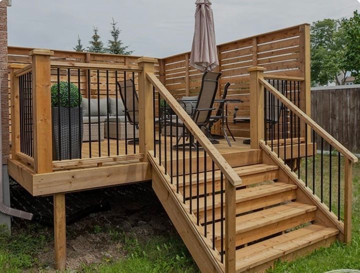 Une terrasse en bois avec des rambardes et des escaliers en métal noir, comprenant des sièges extérieurs et un paravent pour préserver l'intimité, dans un jardin.