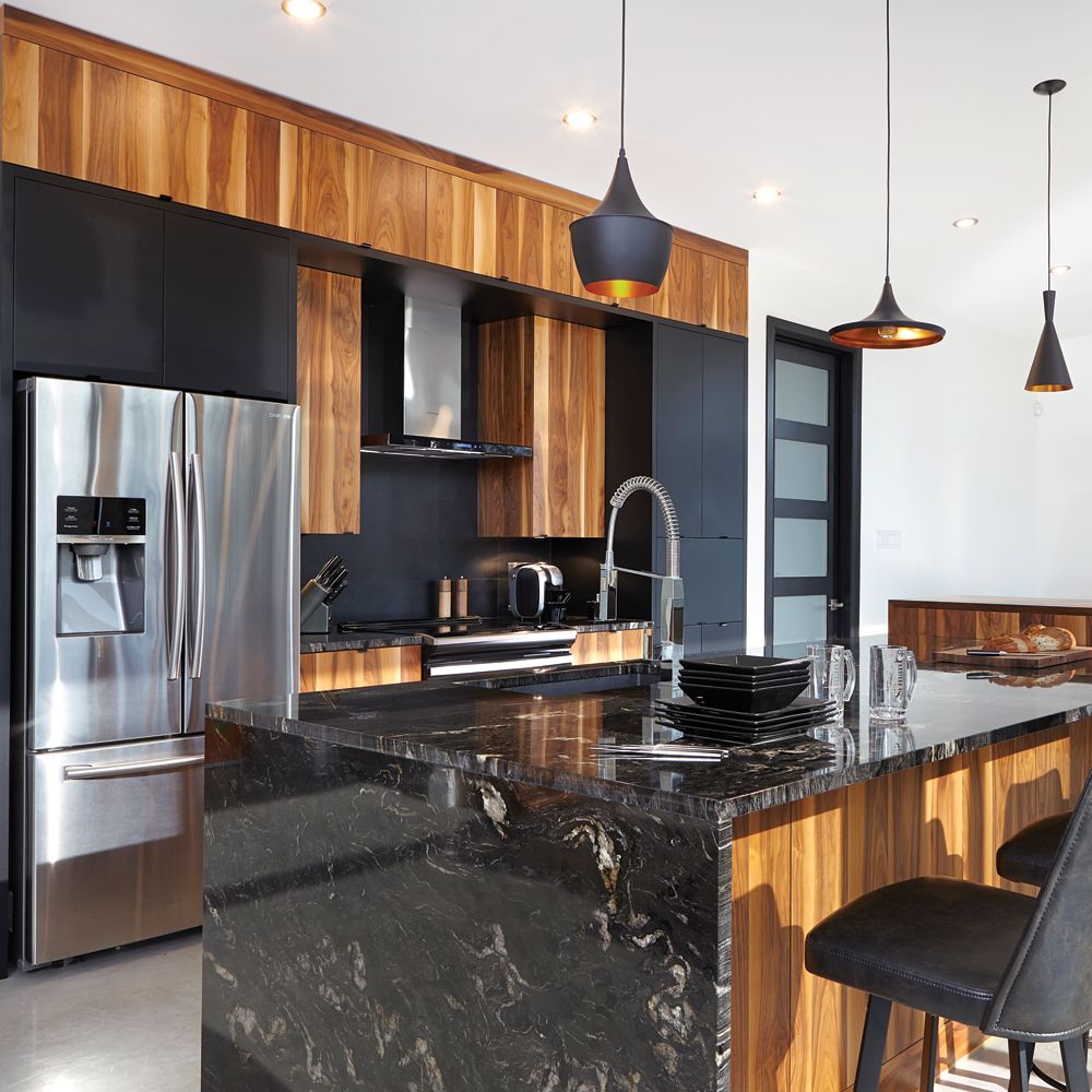 Cuisine moderne avec placards noirs, éléments en bois, îlot en marbre noir, réfrigérateur en acier inoxydable et suspensions lumineuses.