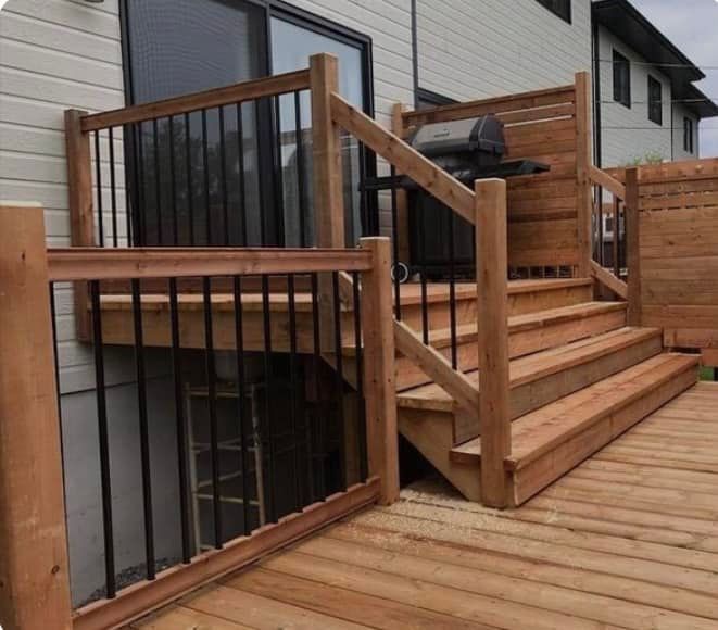 Une terrasse en bois avec escalier et brise-vue jouxte une maison dotée d'une porte coulissante en verre et de balustrades en métal noir.