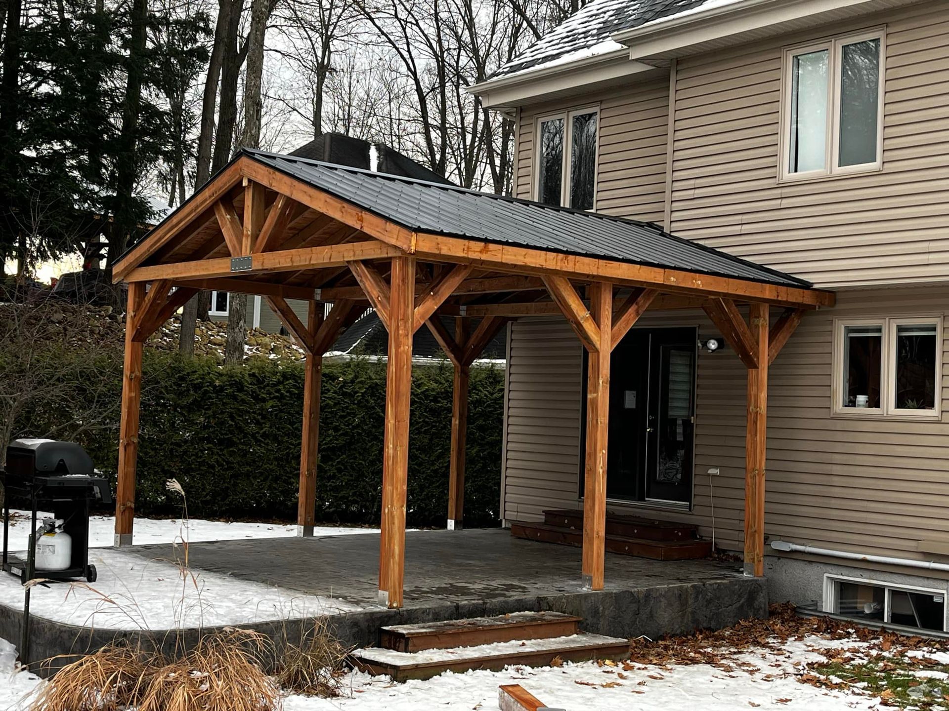 Une pergola en bois, adossée à une maison beige, surplombe une terrasse en pierre par une journée enneigée.
