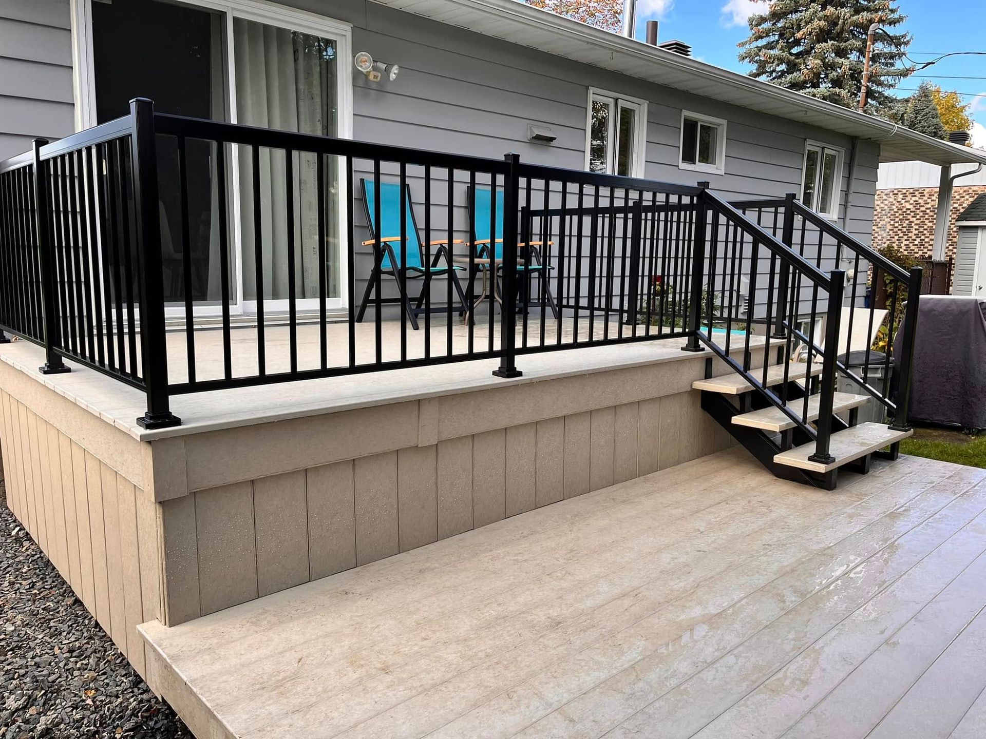 Une terrasse surélevée beige avec des rambardes et un escalier noirs, adossée à une façade de maison gris clair.