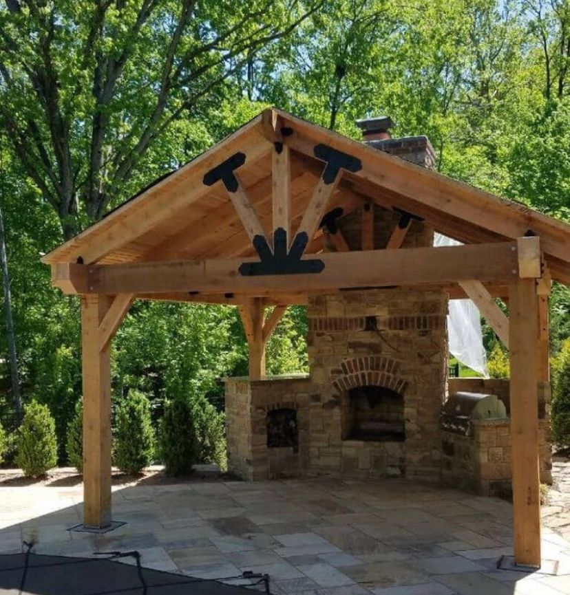 Un pavillon extérieur à ossature bois avec une cheminée en pierre et un barbecue, entouré d'arbres par une journée ensoleillée.