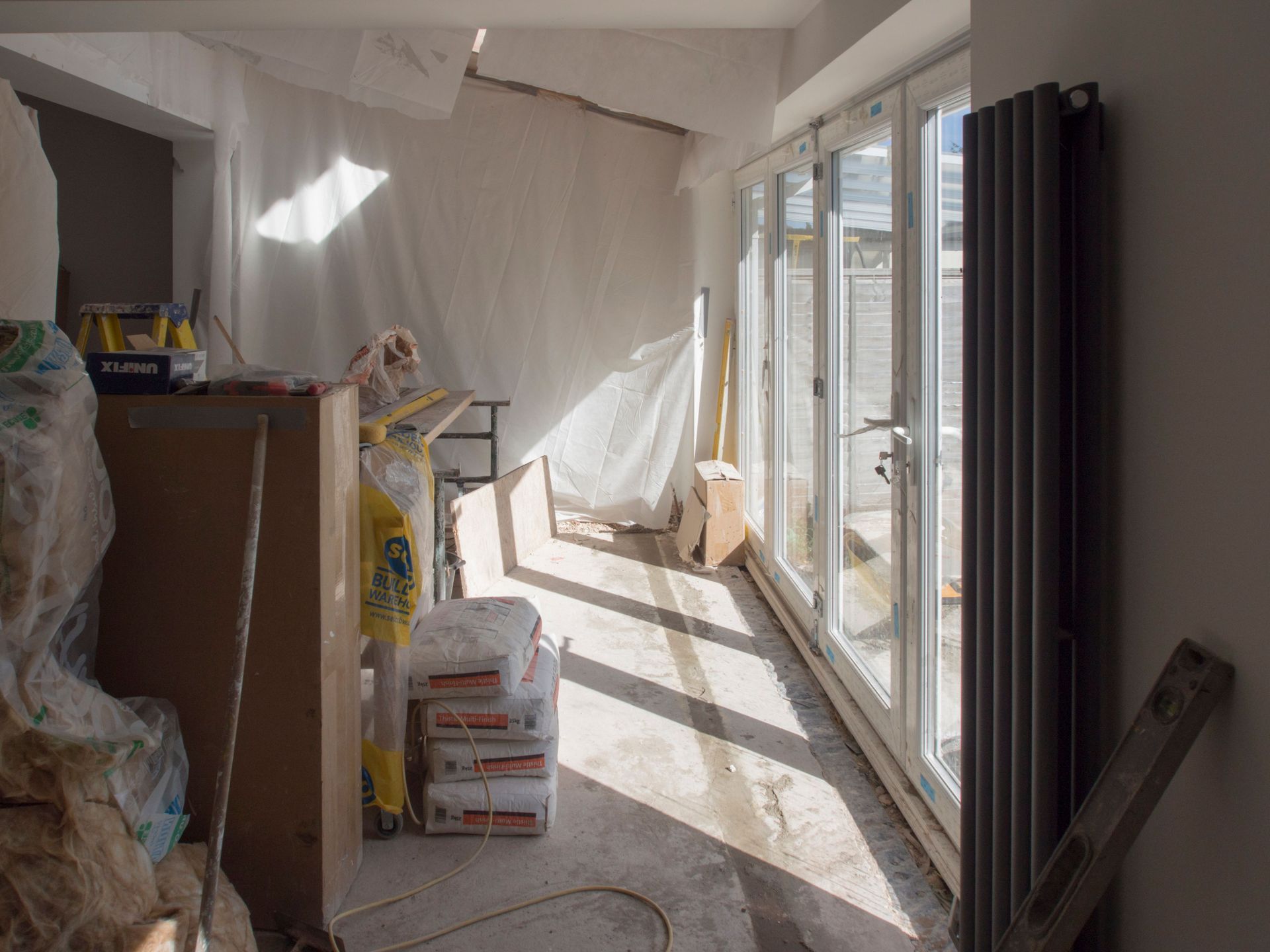 Une extension de maison en forme de L, avec de grandes portes coulissantes en verre, une charpente en bois et des murs extérieurs blancs, est en construction.