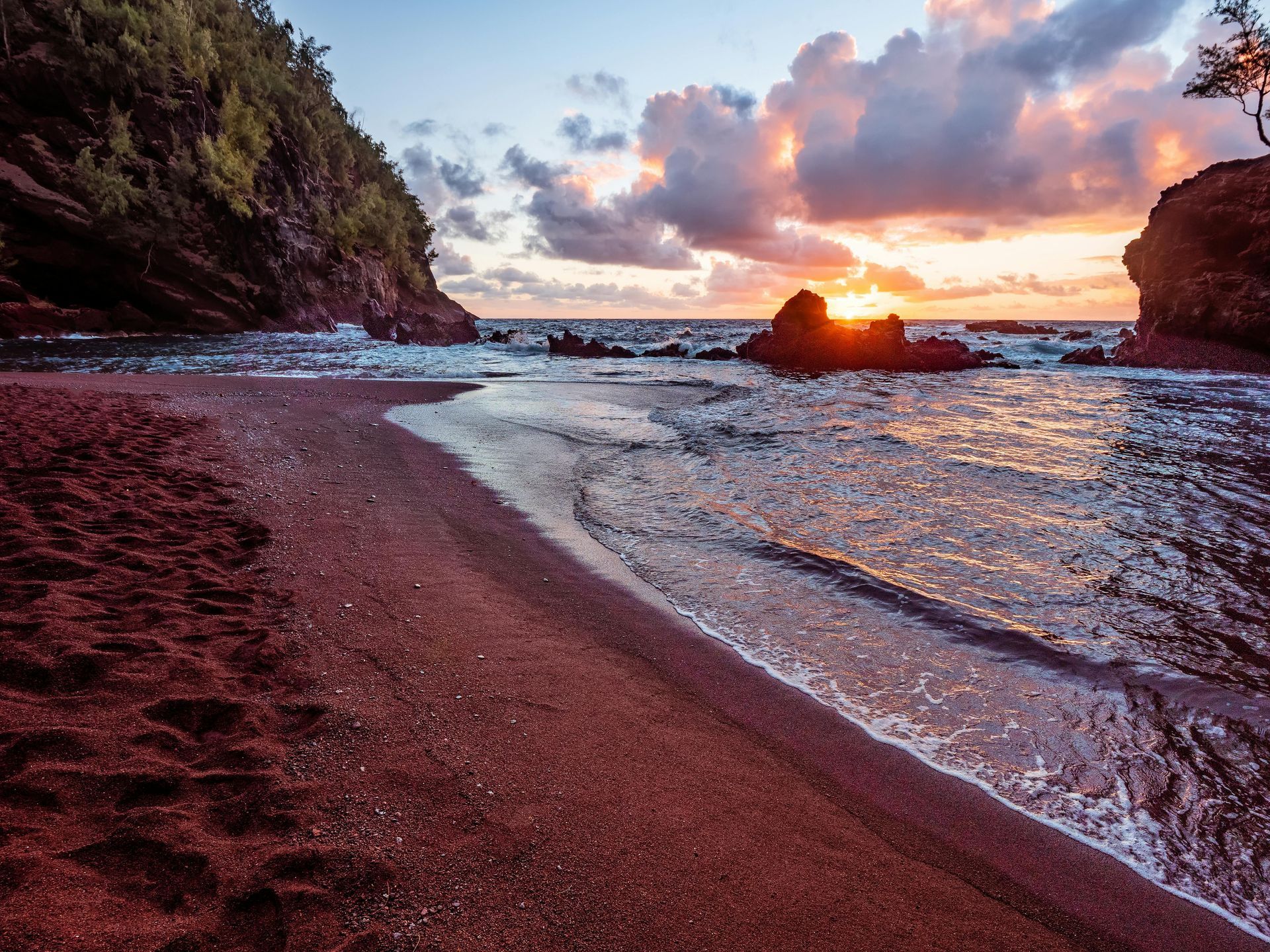Red sand beach at sunset with ocean waves and colorful sky.