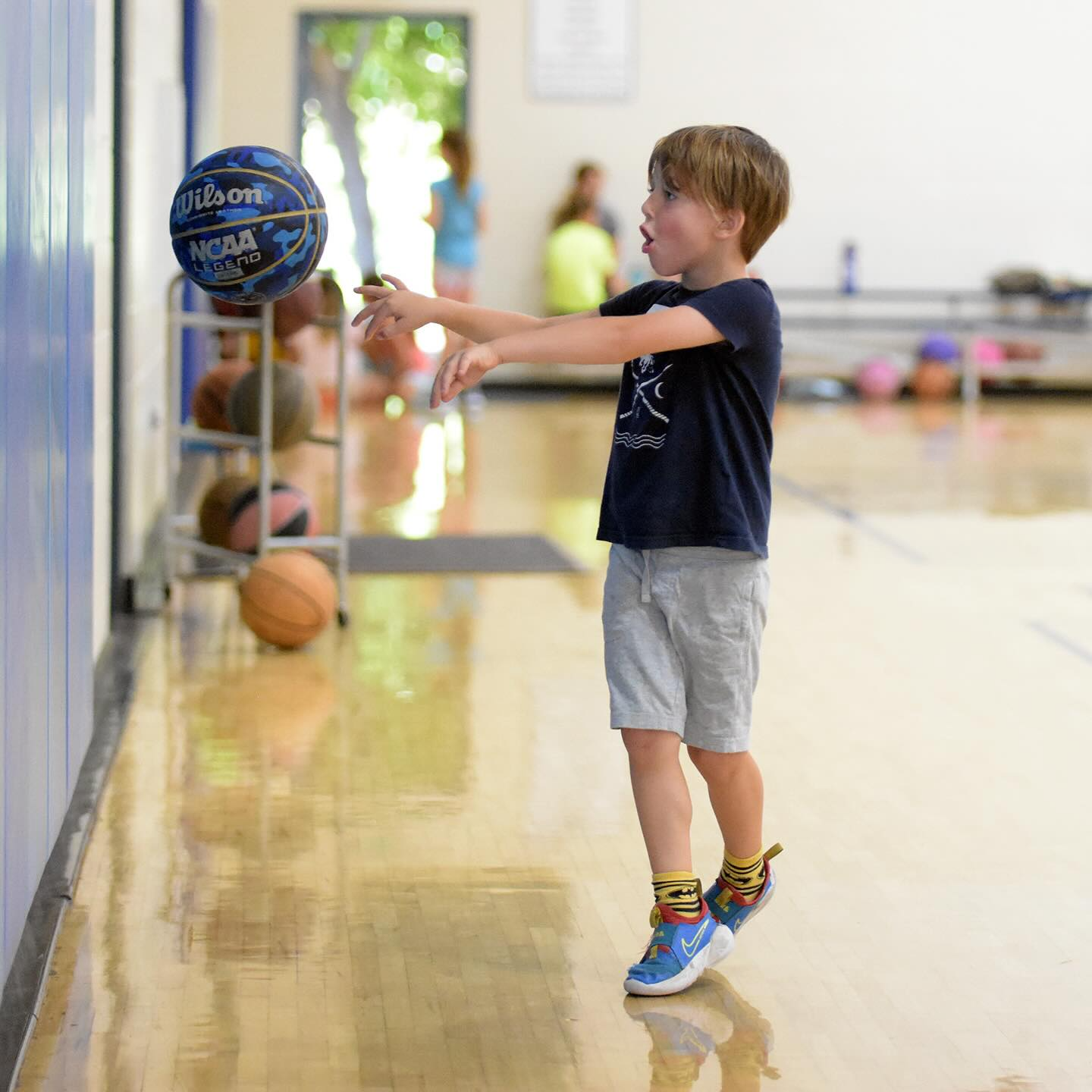 Children playing basketball in a gym, one dribbling while others watch on the court.