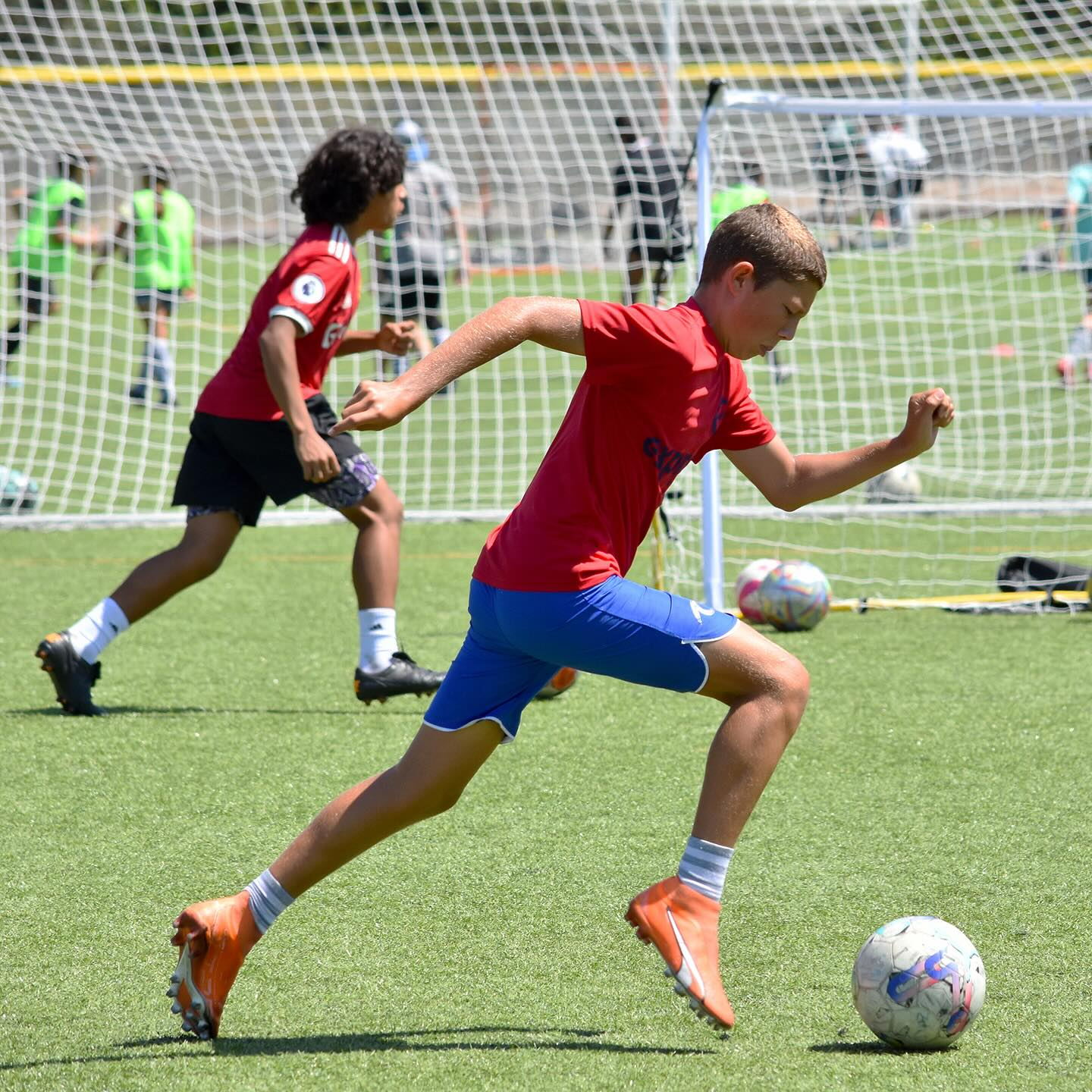 Two soccer players dribble on a grassy field, one in red and white, the other in gray and yellow.