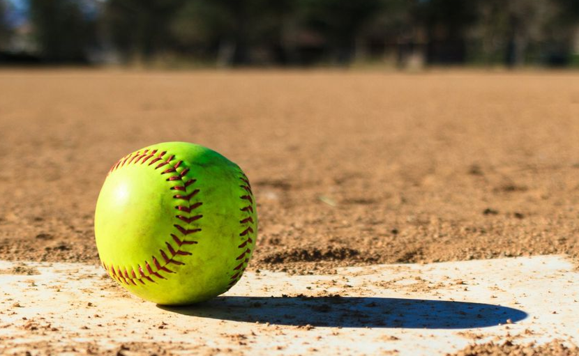 A yellow softball on a dirt field near home plate, with a blurred outfield in the background.