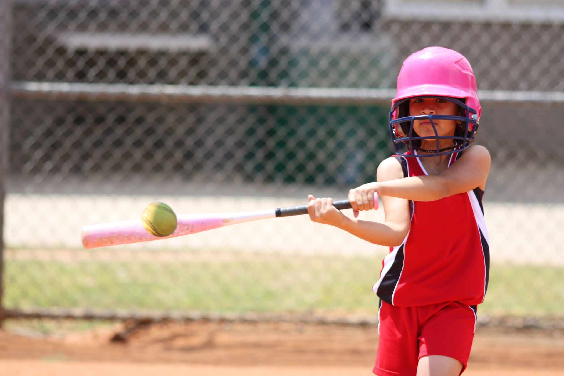 Child in pink helmet and red uniform swings a bat at a ball on a baseball field