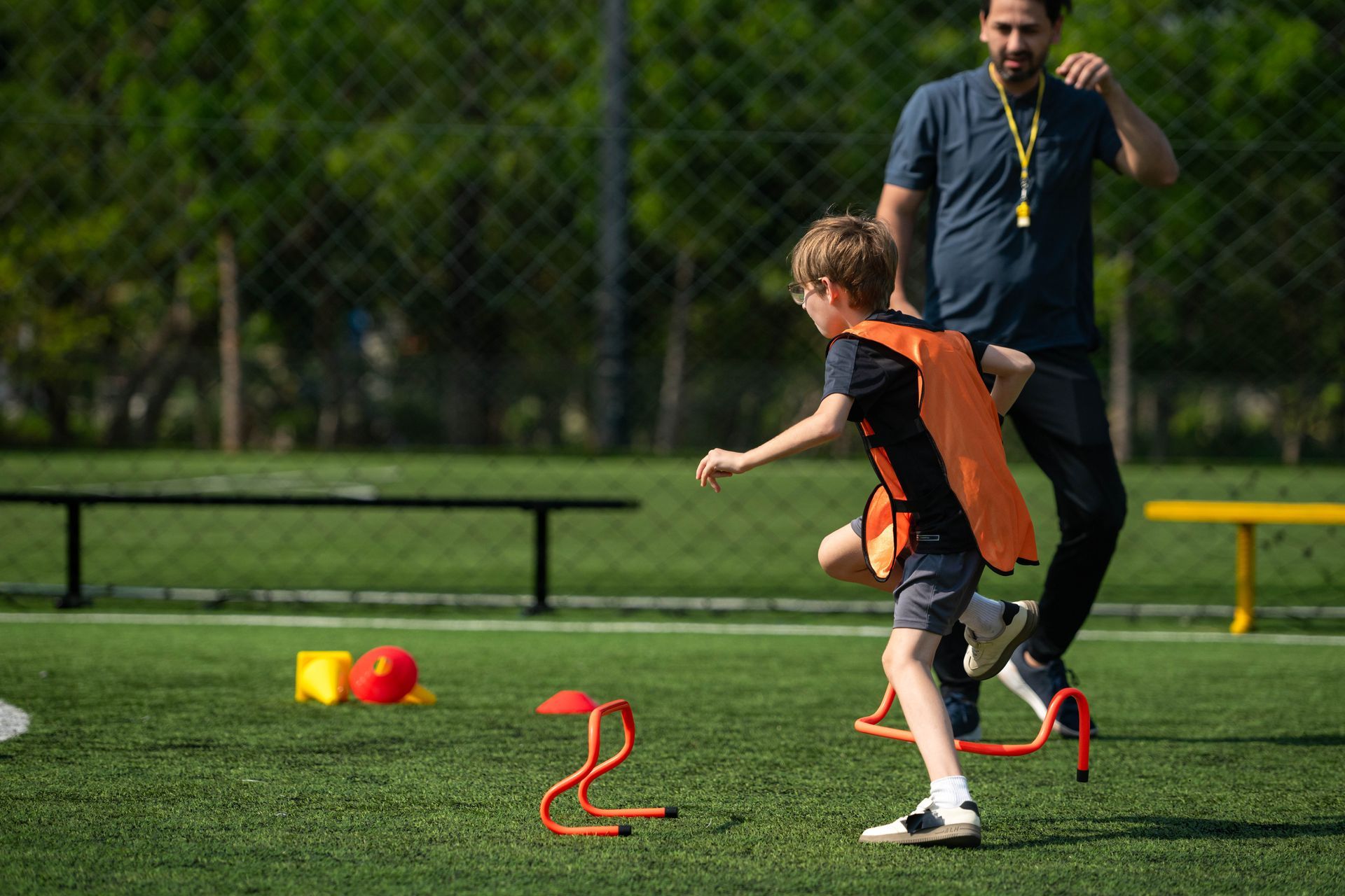 Child in an orange vest runs an agility drill on a grassy field while a coach watches nearby