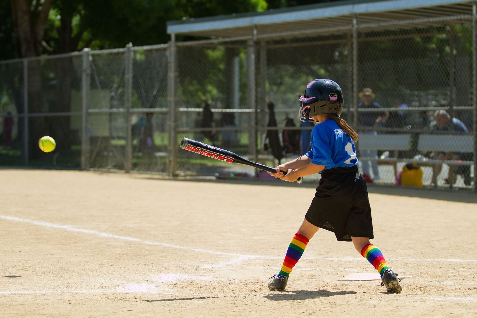 Child in blue batting at a softball on a sunny field, with rainbow socks and a helmet