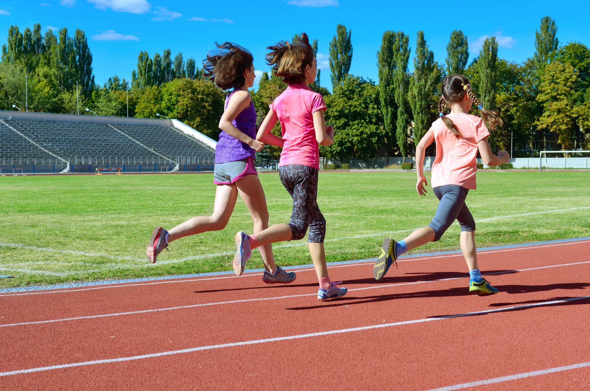 Three runners on a track, seen from behind, jogging beside a green field and trees.