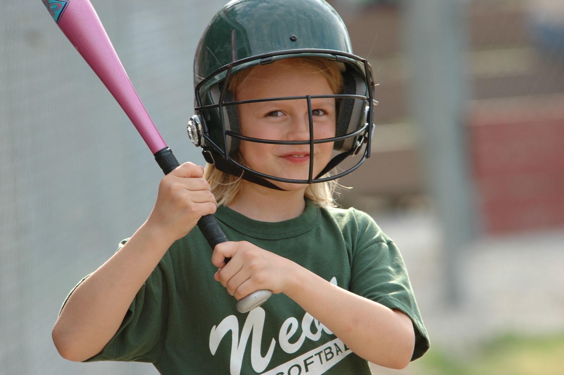 Young baseball player in a green helmet holding a pink bat and smiling in a batting stance