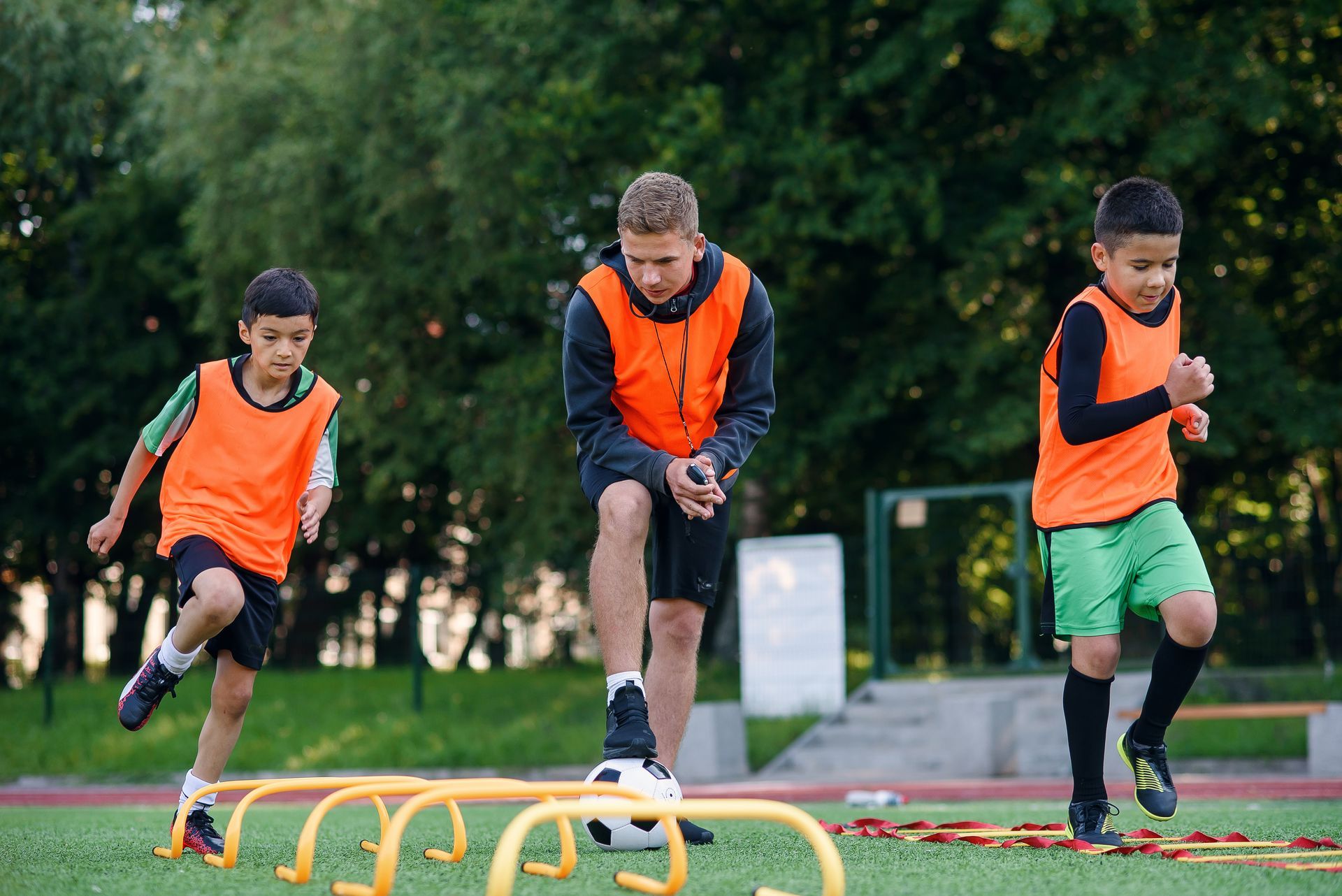 Three children in orange vests doing agility drills over hurdles on a field