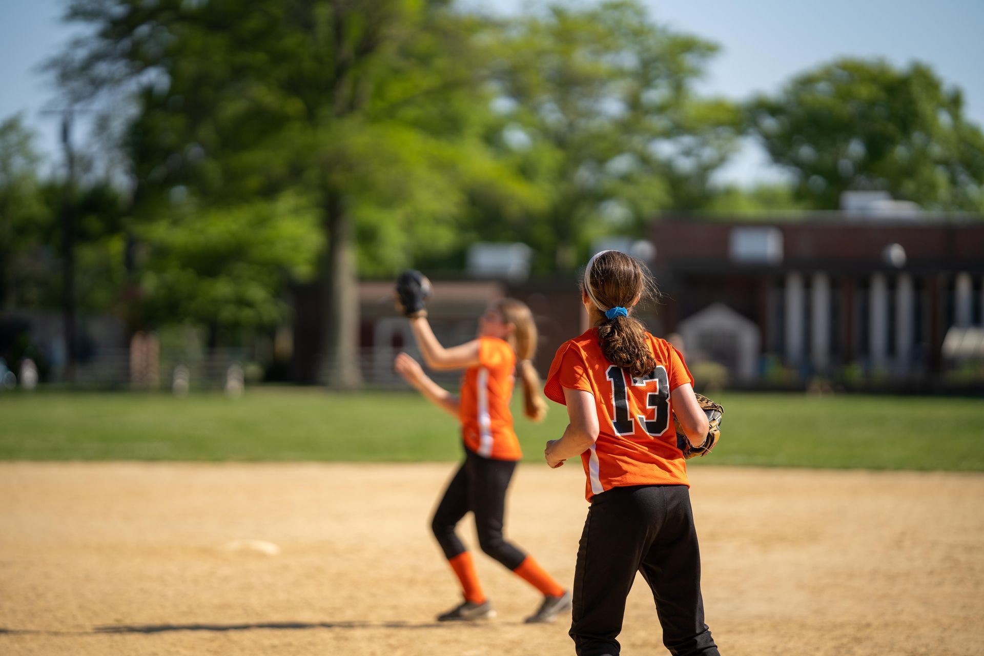 Two softball players in orange jerseys on a field, one catching a ball in the foreground.