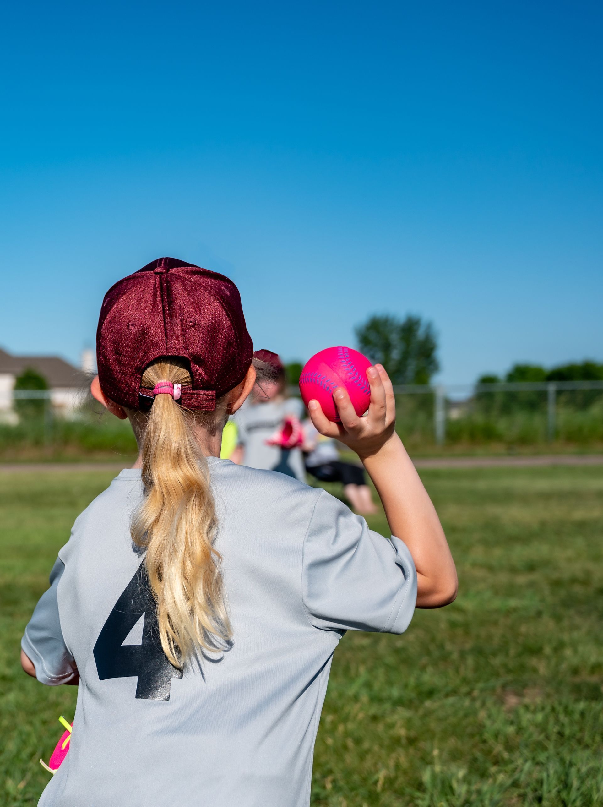 Softball player in a gray jersey and red cap holds a bright pink ball on a sunny field.