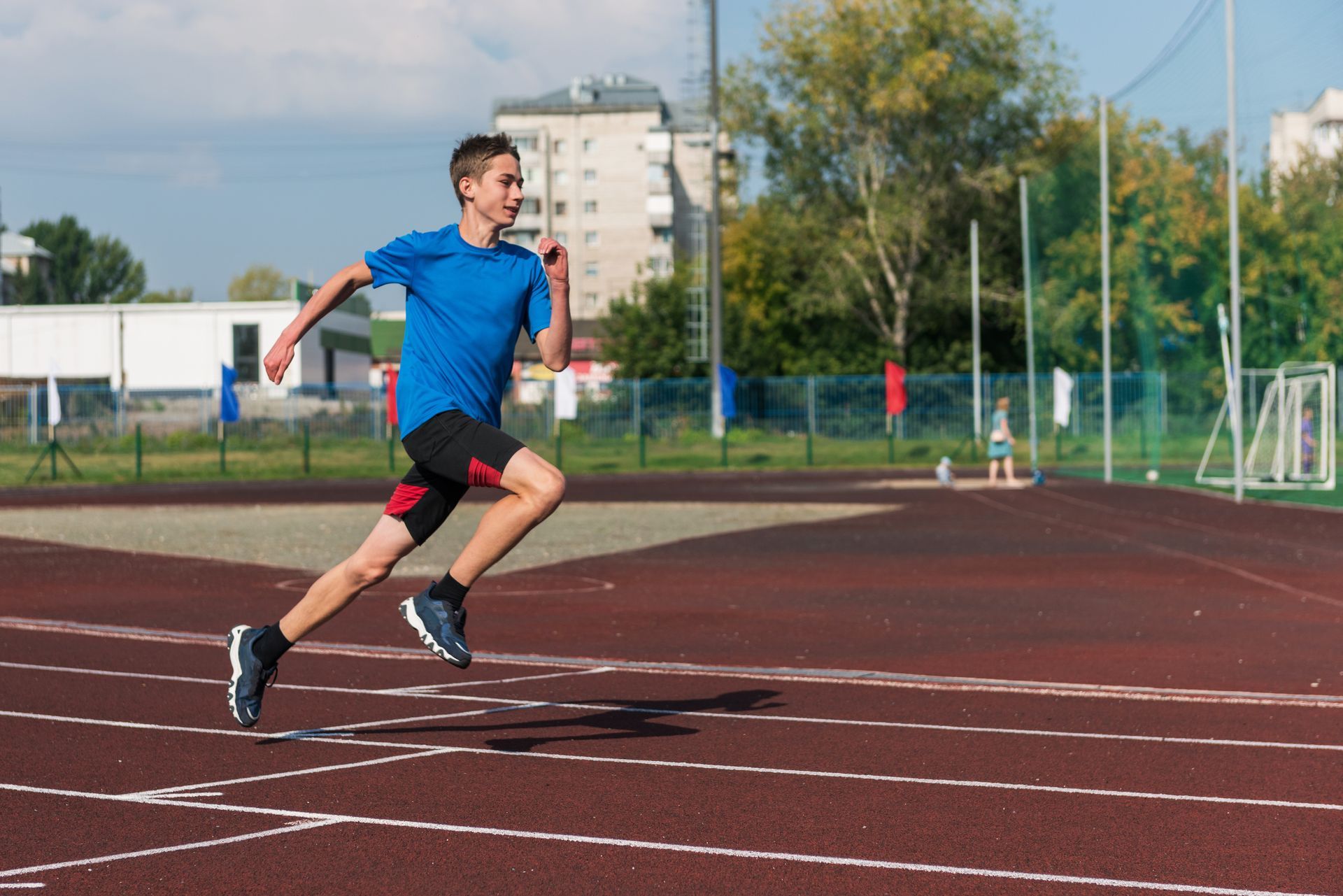 Runner in a blue shirt sprinting on a track outdoors, with a stadium and trees in the background.