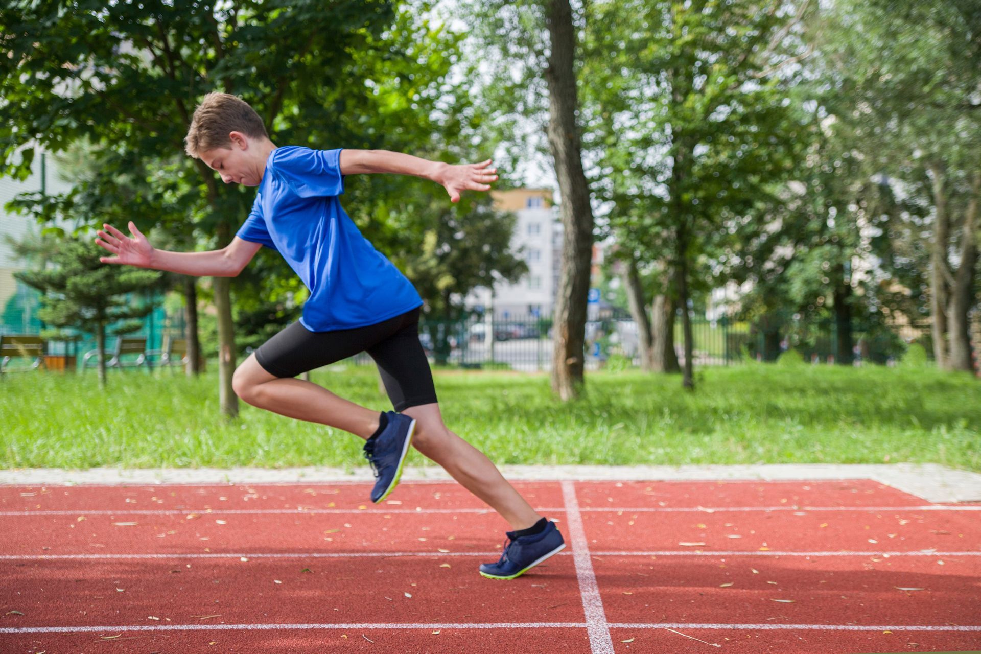 Runner in blue sprinting on a red track, mid-stride with arms outstretched in a park.