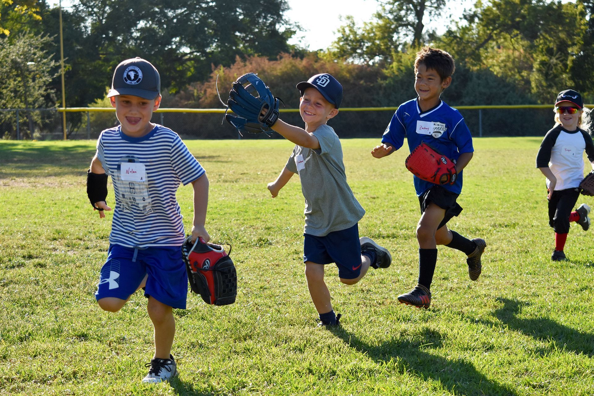 Children playing baseball on a sunny grassy field, with one running toward the camera.