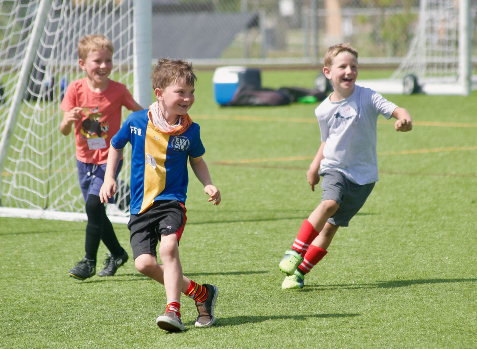 Children playing soccer on a grassy field, running toward a goal in bright daylight.
