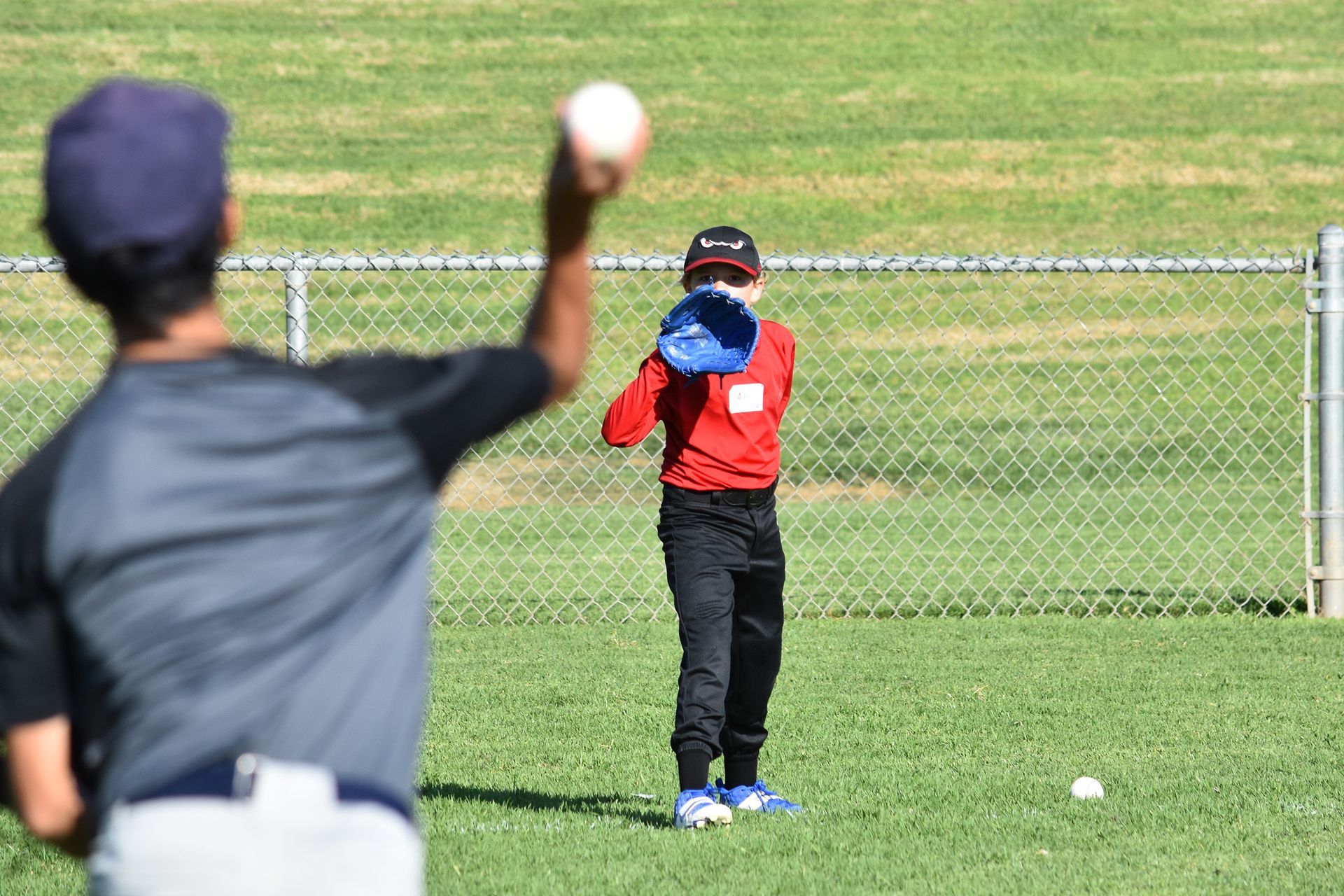 Youth baseball practice: coach throws a ball toward a child in a red jacket on a grassy field.