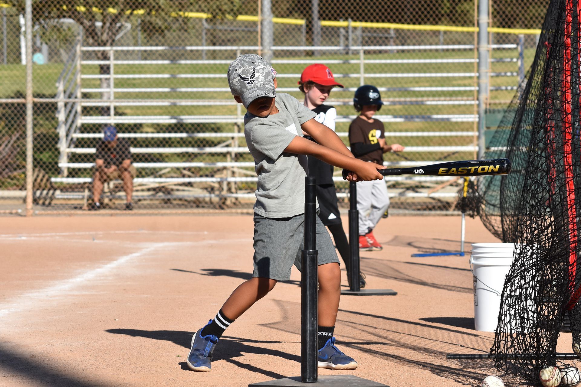 Youth baseball player swinging a bat in a batting cage, with teammates watching nearby.