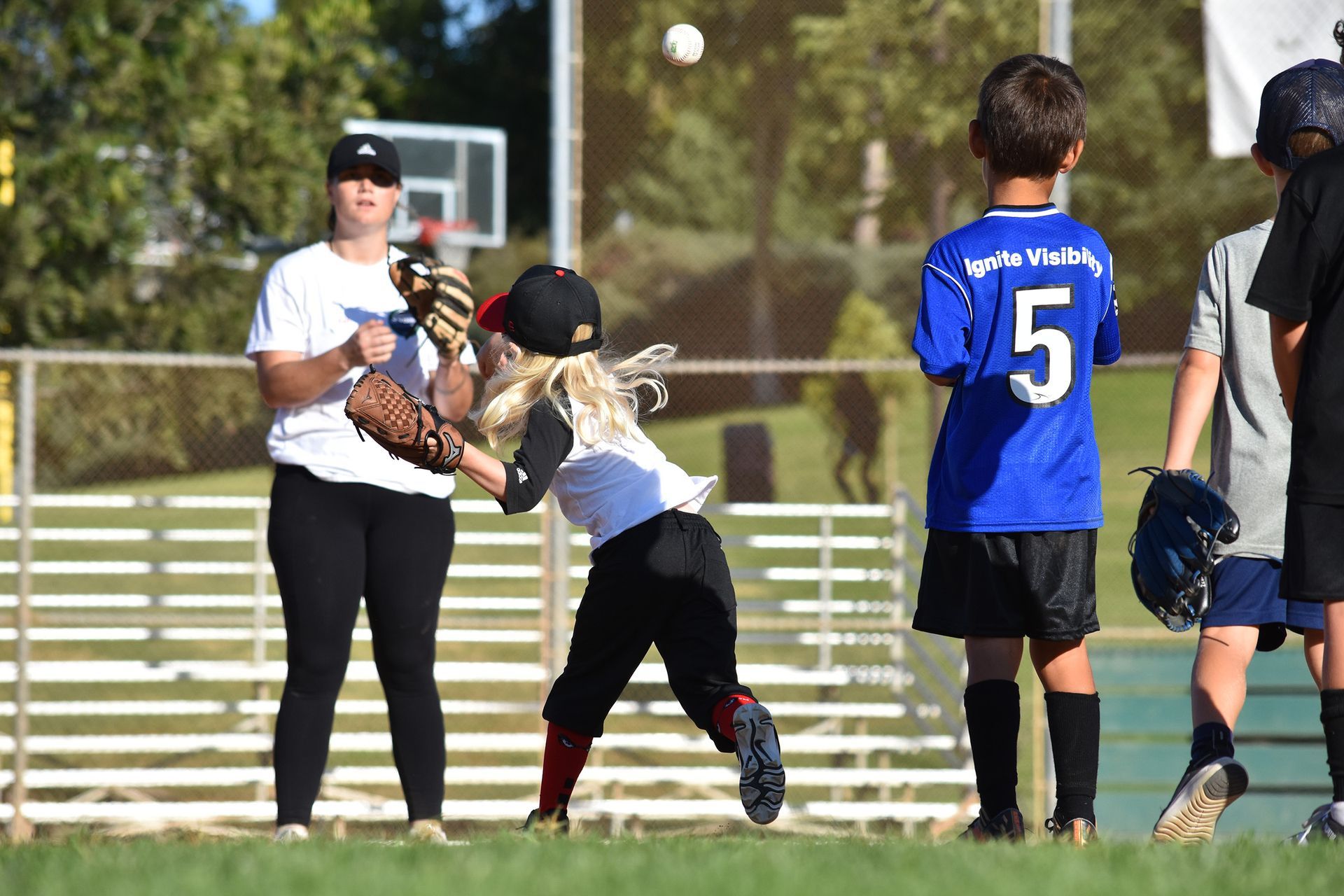 Two baseball players practice batting at a fenced field, one swinging with a coach nearby.