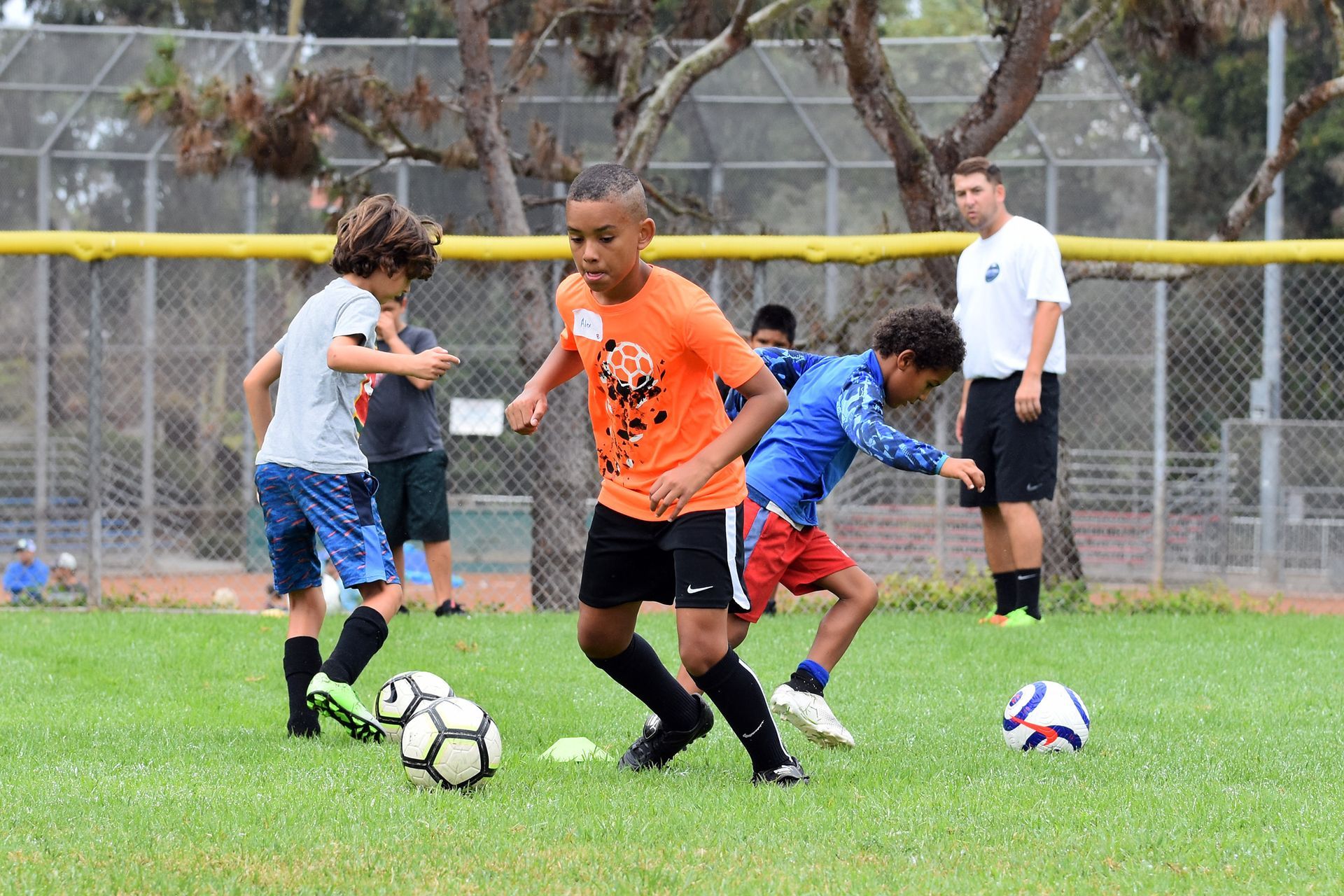 Children playing soccer on a grassy field, with a coach in the background.