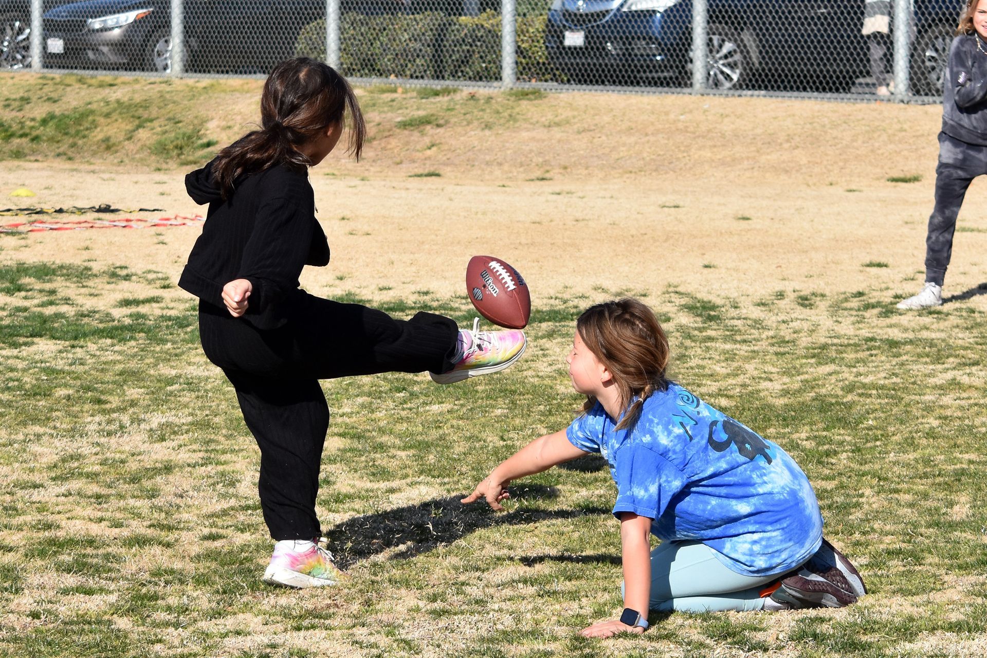 Two children in a field play a game, one kicking a football while another crouches nearby.