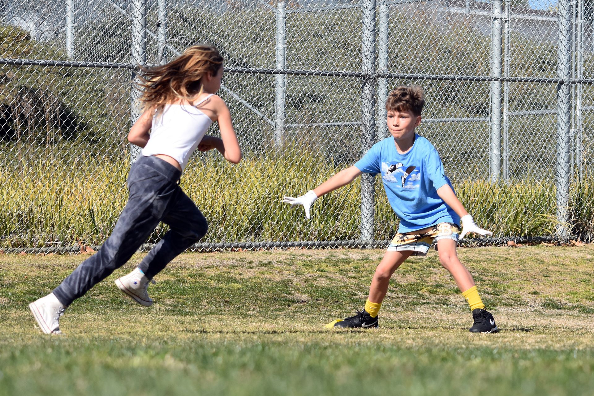 Two people running and reaching in a grassy field near a chain-link fence