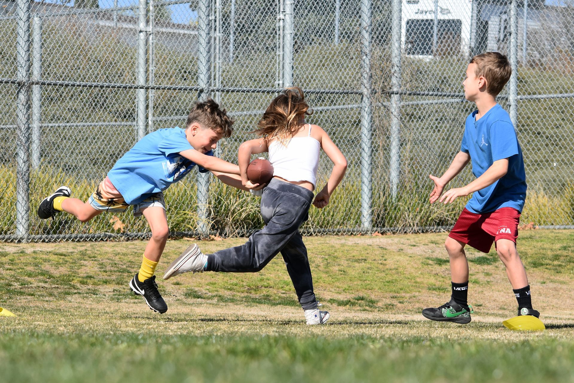 Kids playing tag on a grassy field near a chain-link fence, one boy lunging toward a runner in the center