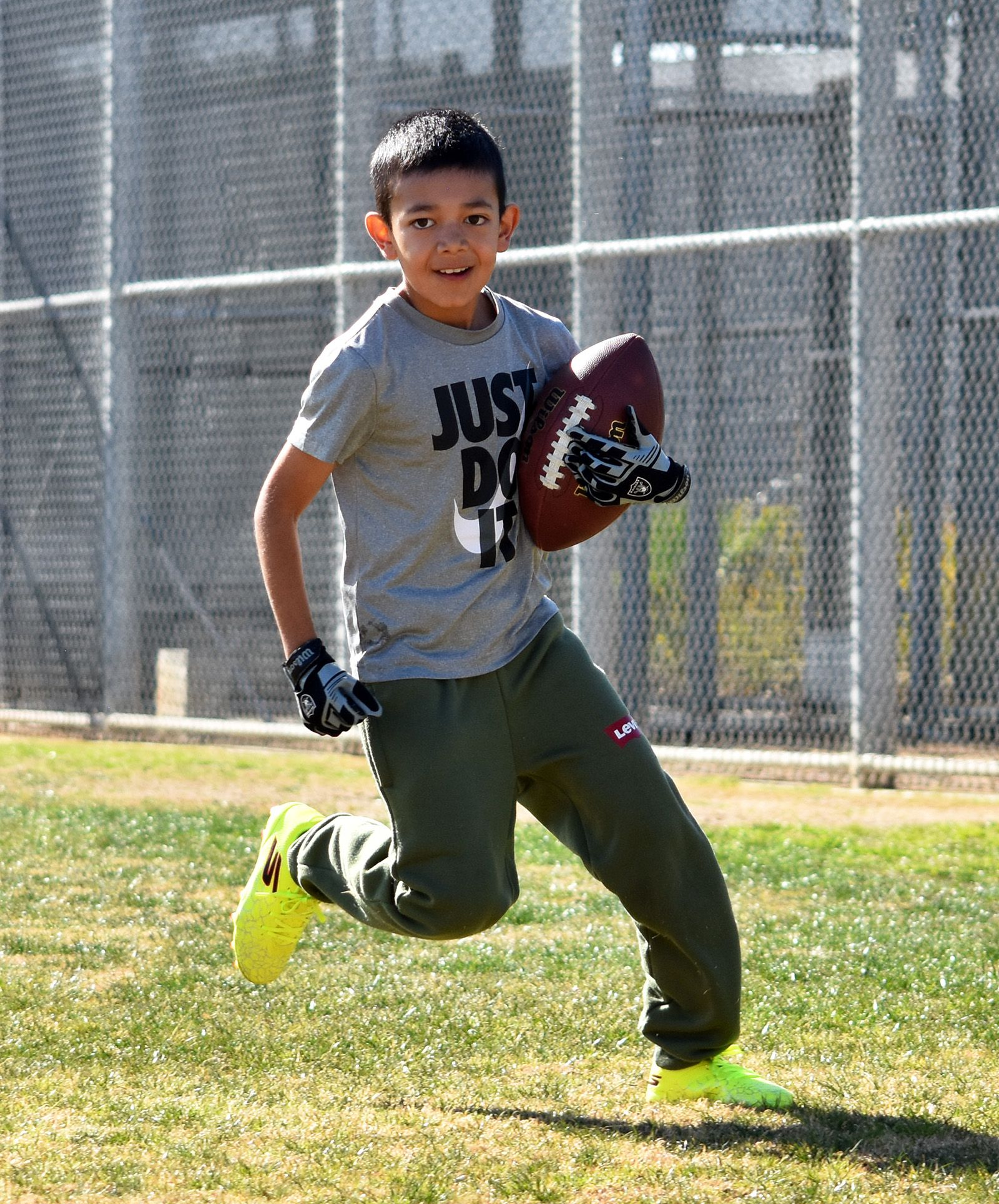 Young athlete running with a football on a grassy field near a chain-link fence