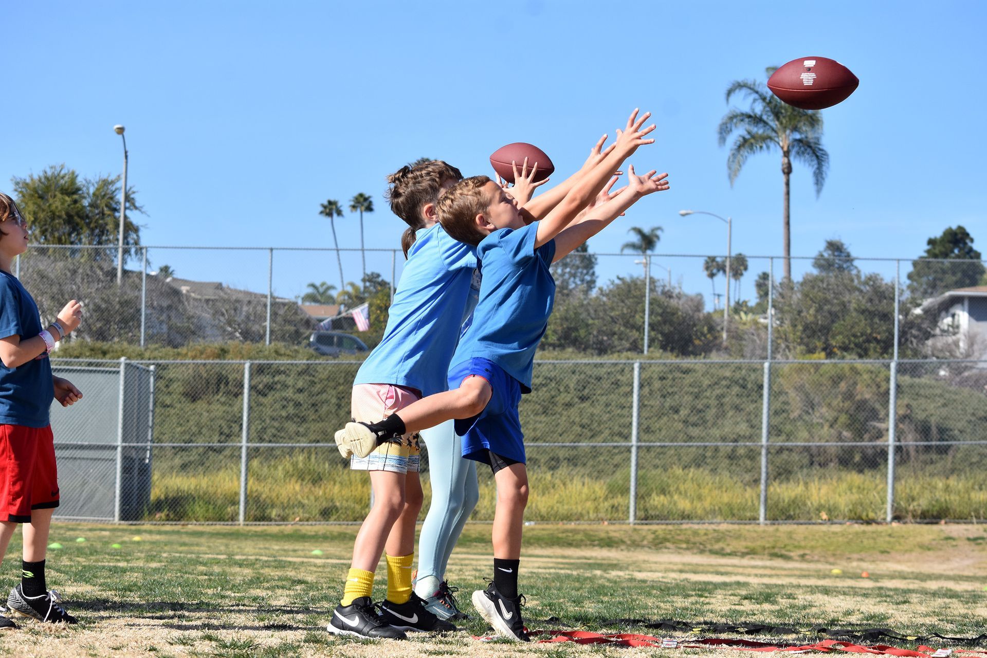 Three players in blue reach up for a football during an outdoor practice drill on a field.