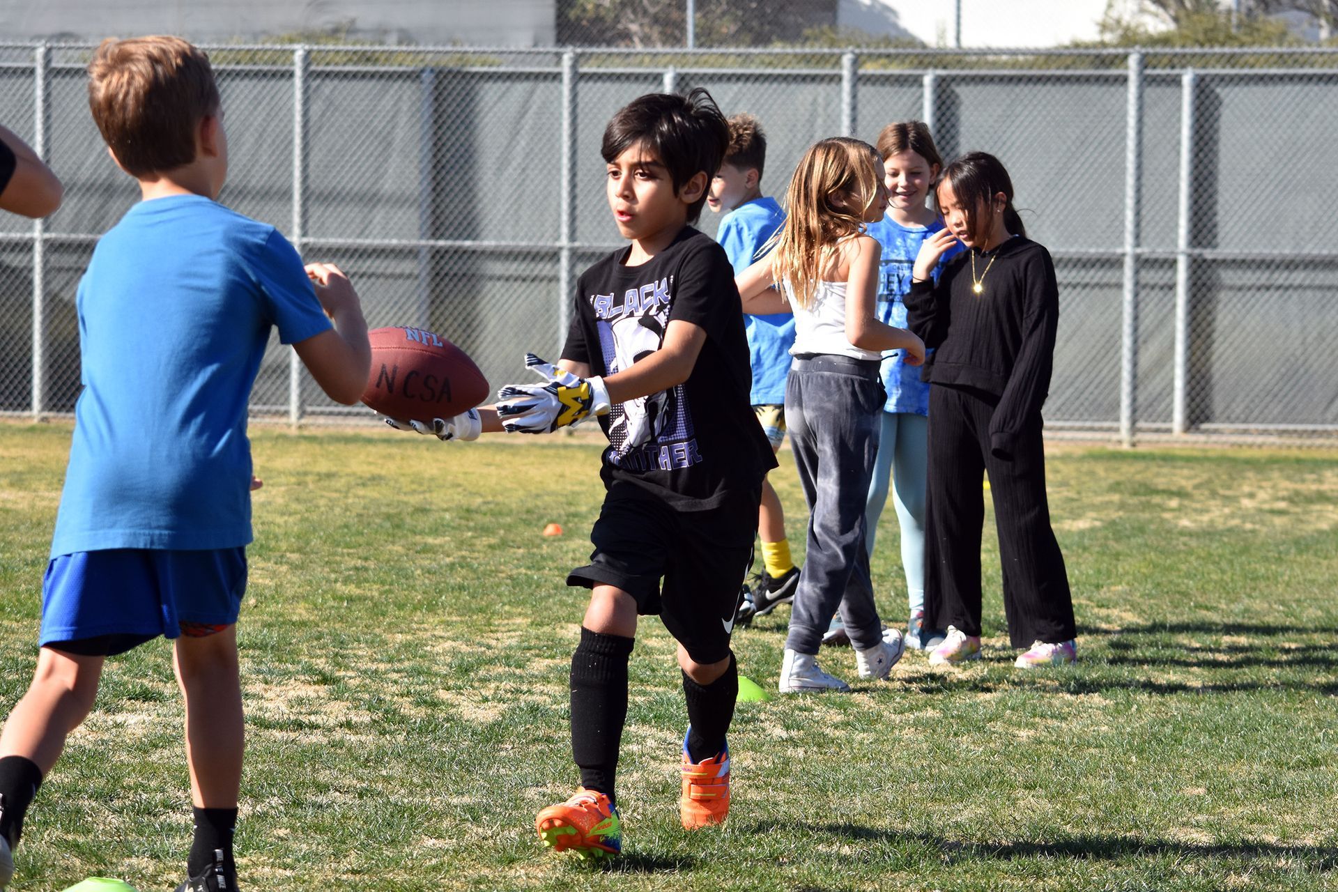 Children playing soccer on a grassy field, one passing a ball to another, with spectators near a fence.