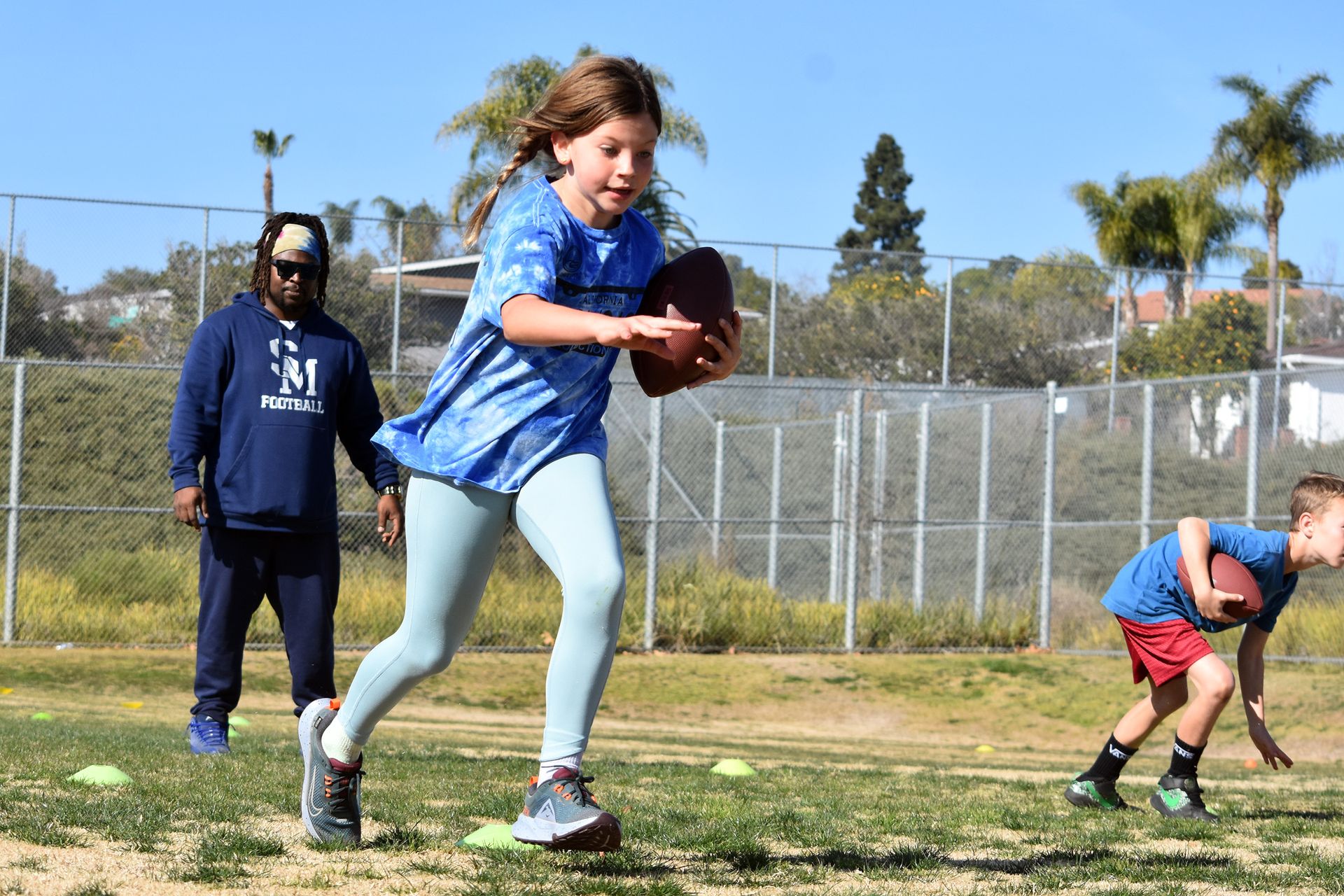 Girl running with a football on a grassy field while two others stand nearby, with a chain-link fence and trees behind.