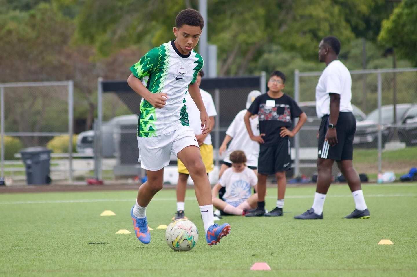 Soccer player dribbling a ball on a field during practice, with other players and cones in the background