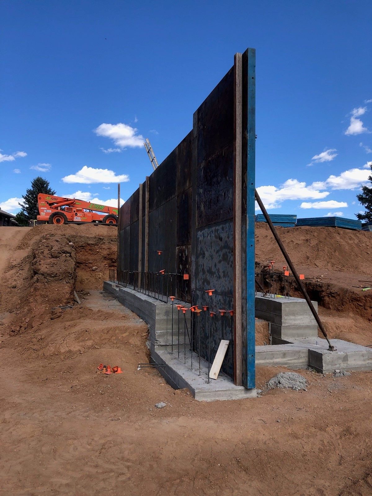 A tall, dark concrete formwork panel stands on a newly poured concrete footing at a construction site under a blue sky.